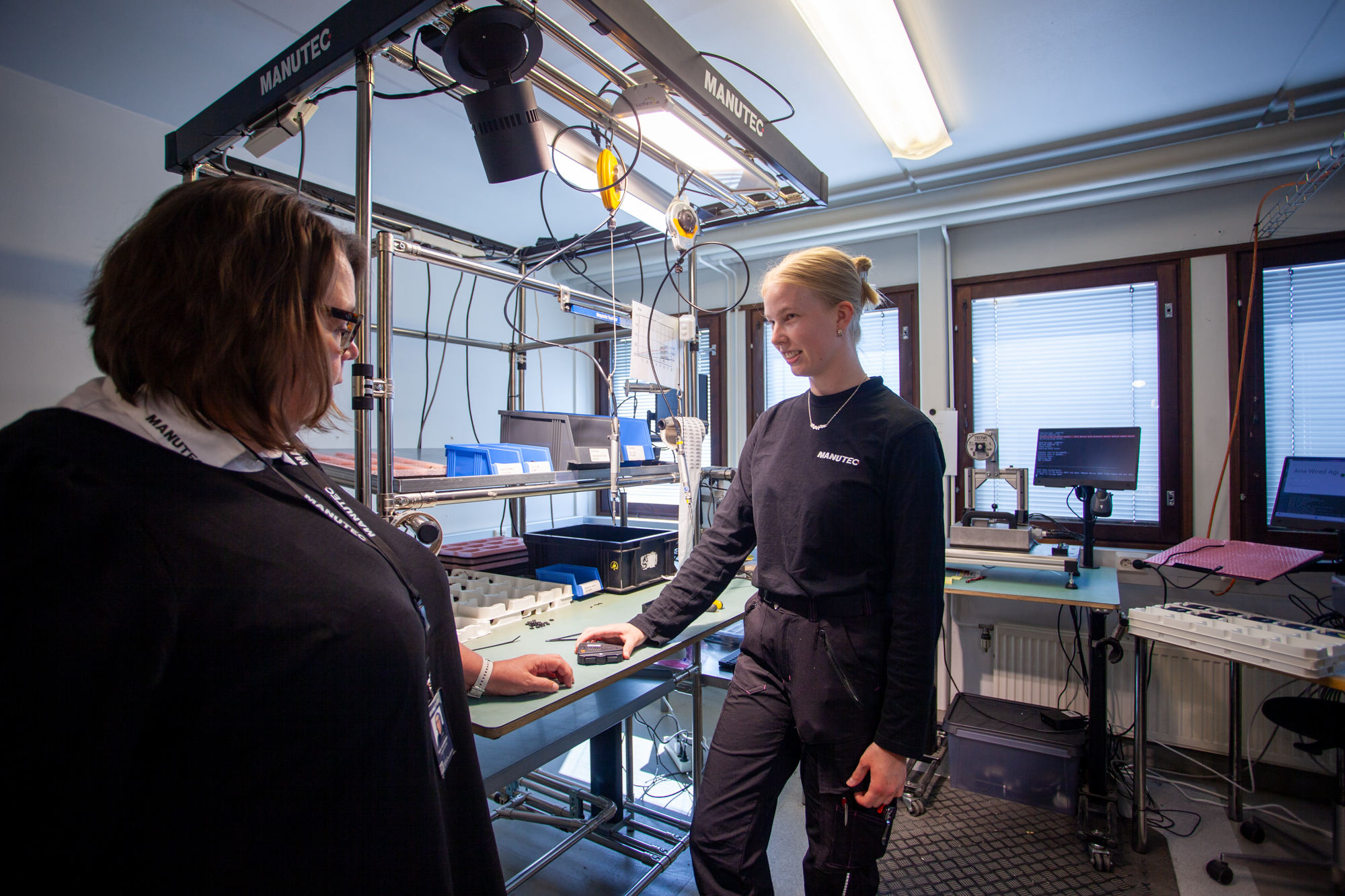 Manutecin toimitusjohtaja Kirsi Junnilainen keskustelee työntekijä Nella Saukkion kanssa yrityksen kokoonpanotilassa. Taustalla näkyy Manutecin tuotantolaitteet ja työpiste.
