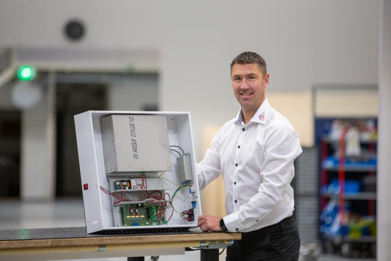 Salo Industry Hubin myyntijohtaja Kristian Johansson katsoo hymyillen kameraan ja esittelee vasemalla puolellaan pöydällä olevaa elektroniikkalaitettalaitetta.